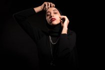 Приваблива молода ісламська жінка, одягнена в чорне вбрання і хіджаб торкається обличчя м'яко і дивиться на камеру — стокове фото