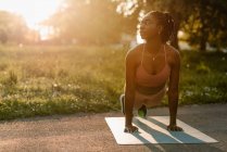 Подходящая афроамериканская спортсменка балансирует в положении доски, выполняя упражнения абс в парке на закате — стоковое фото