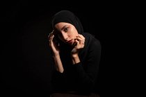 Attraktive junge Islamistin in schwarzem Outfit und Hijab, die das Gesicht sanft berührt und in die Kamera blickt — Stockfoto