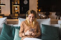 Joyful female sitting on sofa and enjoying music in headphones while looking at screen of smartphone — Stock Photo