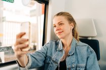 Giovane donna bionda in giacca di jeans che ascolta musica e si fa selfie con il cellulare mentre guida in treno seduta vicino al finestrino — Foto stock