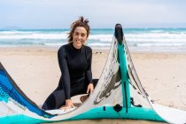 Жінка-кіттер у гідрокостюмі встановлює надувний повітряний змій на піщаному узбережжі океану з рюкзаком та спорядженням на дошці для кіоску — стокове фото
