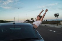 Молода жінка в повсякденному одязі та головах з американським прапором, що виходить з вікна автомобіля і піднімає руки, насолоджуючись свободою під час подорожі на заході сонця. — стокове фото