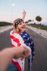 Delighted American female wrapped in USA flag holding hands with man and walking along road while looking at camera — Stock Photo