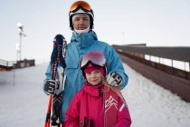 Glad father and daughter wearing warm sports clothes and helmets standing with skis on snowy hill slope and looking at camera contentedly — Stock Photo