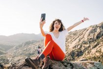 Позитивна молода жінка-мандрівник з кучерявим темним волоссям в повсякденному одязі, сидячи в скелях і посміхаючись, приймаючи селфі на мобільний телефон під час походу в гори в сонячний день — стокове фото