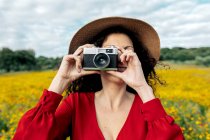 Anónimo sonriente hembra en sombrero tomando fotos en cámara vintage en prado bajo cielo nublado - foto de stock