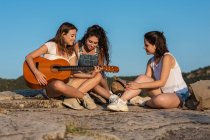 Группа женщин-туристов, сидящих на холме в высокогорье, играющих на акустической гитаре и поющих песни — стоковое фото