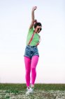 Full body delighted young female wearing shorts and bright pink tights jumping merrily with arm raised on lush grassy glade in nature — Stock Photo