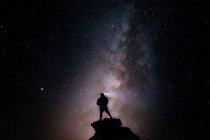 Bajo ángulo de silueta de turista anónimo de pie con antorcha de luz en la cabeza en el acantilado contra el cielo estrellado brillante en la noche - foto de stock