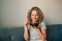 Fröhliche Frau in häuslicher Kleidung genießt leckeres Essen in einer Schüssel, während sie auf einem bequemen Sofa sitzt, die Beine mit einem Lächeln gekreuzt — Stockfoto