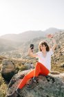 Позитивная молодая женщина-путешественница с кудрявыми темными волосами в повседневной одежде сидит в скалах и улыбается, делая селфи на мобильном телефоне во время похода в горы в солнечный день — стоковое фото