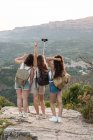 Por trás viajando amigos do sexo feminino com mochilas em pé na colina e tomando auto-tiro no smartphone no fundo da gama de montanhas no verão — Fotografia de Stock