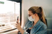 Случайная молодая женщина в джинсовой куртке и медицинской маске фотографируется со смартфоном, сидя на сиденье поезда возле окна — стоковое фото