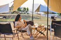 Beautiful ethnic Asian female in sunglasses sitting at table drinking tea while having a relaxing time in camping area during holidays — Stock Photo