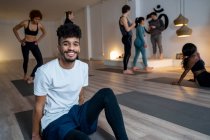 Улыбающийся афроамериканец в активной одежде сидит на коврике и смотрит в камеру после урока йоги на фоне разных людей — стоковое фото