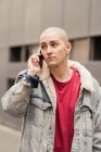 Transgênero pessoa em vestuário casual falando no celular enquanto olha para longe contra o edifício urbano à luz do dia — Fotografia de Stock
