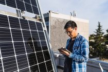 Técnico masculino étnico concentrado en tableta de navegación camisa a cuadros mientras está de pie cerca del panel fotovoltaico ubicado en la granja de energía solar moderna - foto de stock