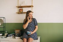 Весела майбутня жінка з чашкою гарячого напою, що говорить на мобільному телефоні, дивлячись в кімнаті — стокове фото