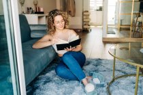 Full body of positive young female with curly hair in casual clothes and slippers sitting on carpet near sofa and reading interesting book during weekend at home — Photo de stock