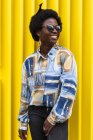 Young African American female with traditional comb on afro hair dressed in stylish colorful informal outfit smiling happily while standing against colorful yellow wall on urban street — Stock Photo