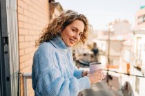 Seitenansicht der zufriedenen jungen Frau mit lockigem Haar im bequemen Kriegspullover, die eine Tasse heißen Kaffee trinkt, während sie sich am sonnigen Morgen auf dem Balkon entspannt — Stockfoto