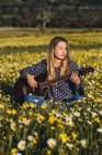 Mujer hipster joven pensativo sentado en un prado en el campo tocando la guitarra durante la luz del sol de verano - foto de stock