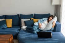 Encantada grávida deitada no sofá confortável e assistindo vídeo engraçado no netbook enquanto relaxa em casa — Fotografia de Stock