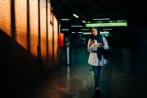 Femme musulmane élégante dans le hijab debout avec café à emporter dans le métro et surfer sur Internet sur téléphone mobile — Photo de stock