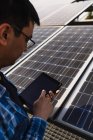 Концентрированный этнический мужчина-техник в клетчатой рубашке просмотра планшета, стоя рядом с фотоэлектрической панели, расположенной в современной солнечной электростанции — стоковое фото