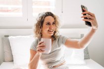 Alegre joven hembra con taza de tomar selfie en el teléfono inteligente mientras está sentado en la cama cómoda - foto de stock