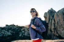 Seitenansicht einer fröhlichen jungen Forscherin in Kapuzenpulli und Sonnenbrille, die lächelt, während sie die Natur beim Trekking im felsigen Gebirgstal an sonnigen Tagen bewundert — Stockfoto