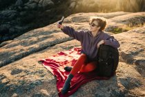 Joyful young female hiker with curly hair in casual outfit and sunglasses leaning on backpack and taking selfie with smartphone while relaxing on rocky hill slope on sunny day — Stock Photo