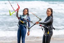Atletas femininas multiétnicas em fatos de mergulho com prancha e barras de controle olhando umas para as outras na costa arenosa contra o oceano espumoso — Fotografia de Stock
