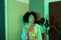 Schwarze Sängerin performt Song gegen Mikrofon mit Popfilter, während sie mit der Hand auf Hüfte und geschlossenen Augen im Tonstudio steht — Stockfoto