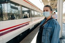 Young female in casual clothes and protective mask standing on railway station and looking away while waiting for train — Stock Photo