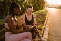 Alegre multirracial atletas do sexo feminino em activewear sentado no banco no parque e usando telefones celulares juntos após o treinamento ao pôr do sol — Fotografia de Stock