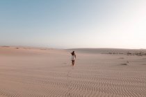 Вид ззаду анонімного туриста-жінки в білій сукні, що йде на ребристий пісок під світлим небом — стокове фото