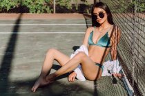 Seitenansicht einer jungen sanften Touristin in Badebekleidung und Sonnenbrille, die im Urlaub mit überkreuzten Beinen hinter Gitterzaun sitzt — Stockfoto