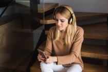 Fröhliche Frau mit Kopfhörer genießt Lieder, während sie zu Hause auf der Treppe sitzt und auf dem Smartphone surft — Stockfoto