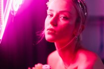 Attraktive junge barbusige Frau in weißen Hosen mit einem Strauß frischer bunter Blumen sitzt auf einem Hocker vor heller Neonbeschriftung im dunklen Studio — Stockfoto