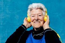 Entzückte ältere Frau mit grauen Haaren und gelben Kopfhörern genießt Lieder, während sie im Studio Musik auf blauem Hintergrund hört — Stockfoto