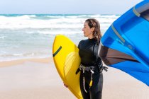 Sportswoman in muta con aquilone gonfiabile passeggiare sulla riva sabbiosa guardando dall'altra parte contro l'oceano tempestoso — Foto stock