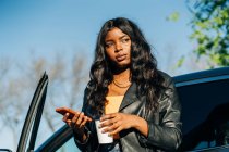 Уверенная афроамериканка со смартфоном и напитком на вынос, стоящая рядом с черным роскошным автомобилем и отворачивающаяся — стоковое фото