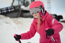 Positivo ragazza carina in rosa caldo activewear occhiali e casco sci accanto pista innevata sulla chiara giornata invernale — Foto stock
