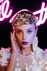 Charming tender young bride in white lace gown and luxurious floral wreath and earrings looking at camera against black background with neon lights — Stock Photo