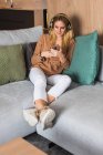 Gioiosa donna seduta sul divano e che si gode la musica in cuffia mentre guarda lo schermo dello smartphone — Foto stock