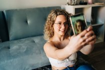 From above of self assured young female millennial with curly blond hair in leather bra an jeans sitting on floor near sofa and taking selfie on smartphone — Foto stock