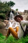 Веселый взрослый турист в повседневной одежде с рюкзаком, сидящим на берегу озера рядом с живописным водопадом при прикосновении и взгляде на камеру — стоковое фото
