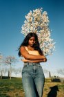 Baixo ângulo da bela fêmea afro-americana em pé no florescente parque primaveril e desfrutando do tempo ensolarado olhando para a câmera — Fotografia de Stock